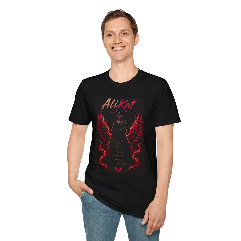 A smiling person wears the AliKat Rising Phoenix Vol. 1 Graphic Tee—a black shirt available in sizes S, M, L, XL, and 2XL—featuring a fiery cat with red wings and the phrase “I rose. I rebuilt. I became unstoppable.” They stand with one hand in their pocket against a white background.