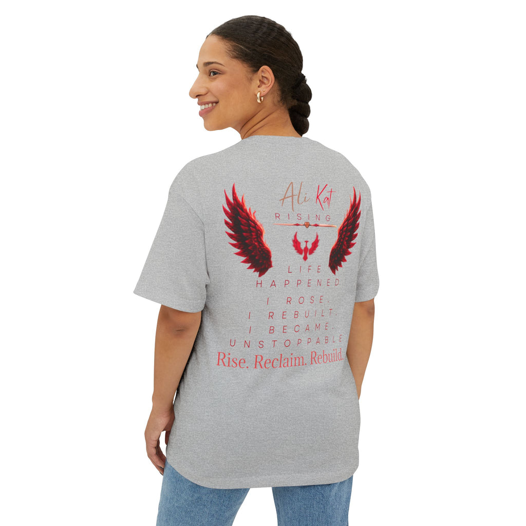 A woman stands smiling, facing away from the camera in a gray, oversized unisex Phoenix Season: Vol. 1 Signature Oversized Tee by AliKat Rising, crafted from premium heavyweight cotton. The back of the shirt features red and black wing graphics with motivational text that reads: Rise. Reclaim. Rebuild.