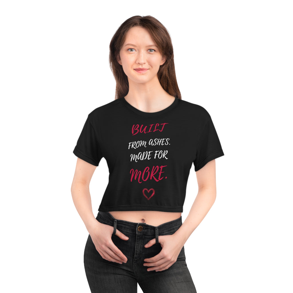 A woman is seen in the AliKat Rising Phoenix Wings Crop Tee — "Built from Ashes, Made for More" Inspirational Women's Top: a black, all-over print crop featuring bold white and red text and a small red heart beneath. This lightweight polyester tee is perfect for standing out as she smiles confidently with her hands in front.