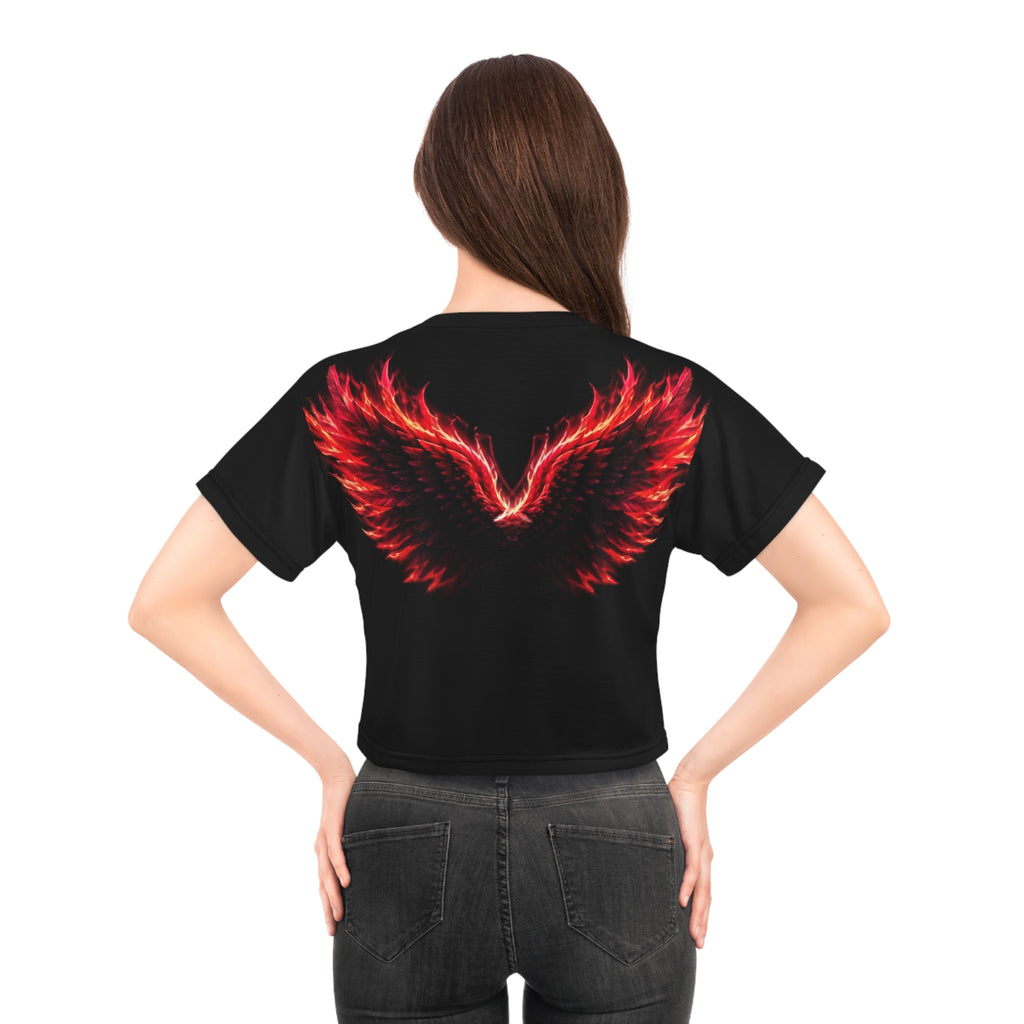 A woman with long brown hair models the AliKat Rising Phoenix Wings Crop Tee—"Built from Ashes, Made for More" Inspirational Women's Top. The lightweight polyester cropped shirt showcases a striking red and orange flaming wings graphic on the back. She pairs it with black jeans and stands with her hands on her hips, facing away.