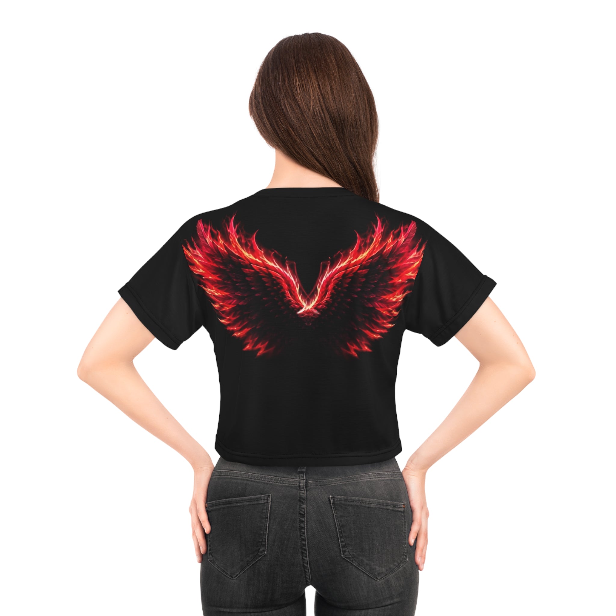 A woman with long brown hair models the AliKat Rising Phoenix Wings Crop Tee—"Built from Ashes, Made for More" Inspirational Women's Top. The lightweight polyester cropped shirt showcases a striking red and orange flaming wings graphic on the back. She pairs it with black jeans and stands with her hands on her hips, facing away.