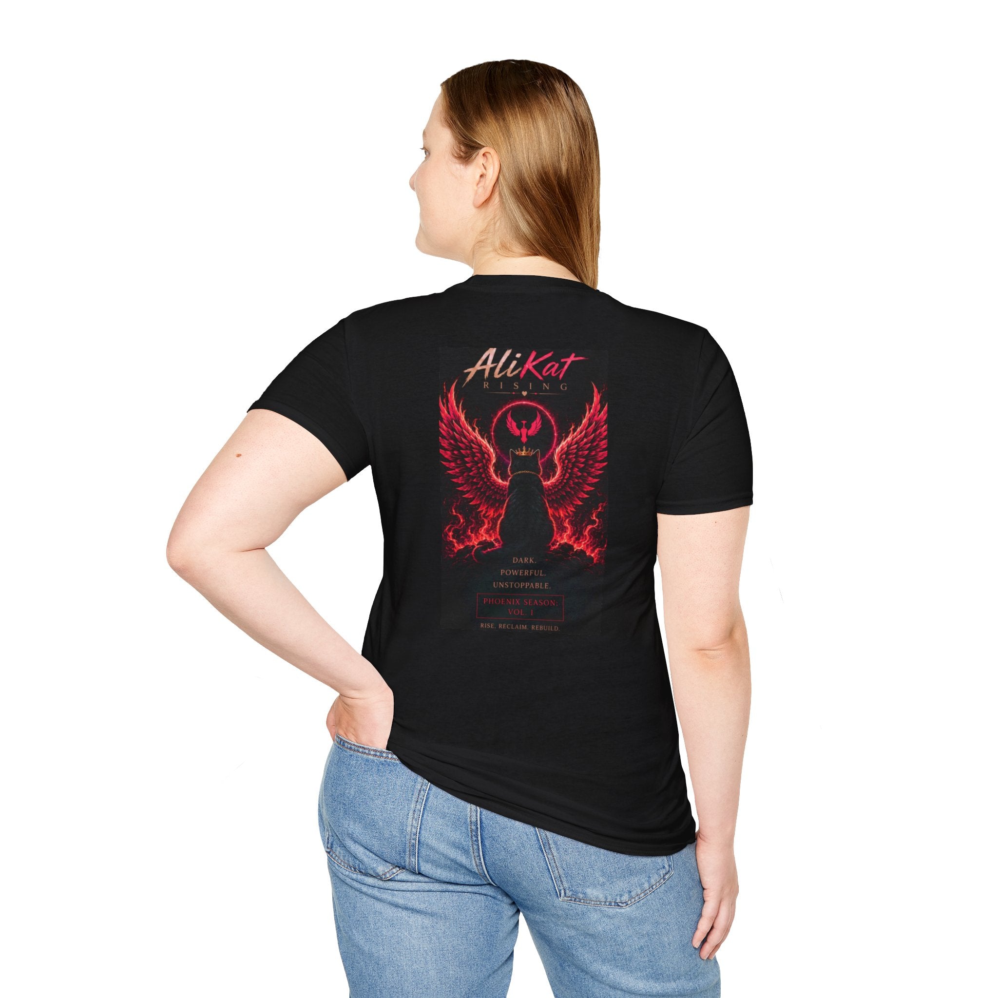 A woman with light brown hair models the Phoenix Vol. 1 Graphic Tee by AliKat Rising—available in S, M, L, XL, and 2XL. The black T-shirt features a striking red winged graphic and the word "AliKat" on the back. She pairs it with blue jeans and poses with her hand on her hip, facing away from the camera.