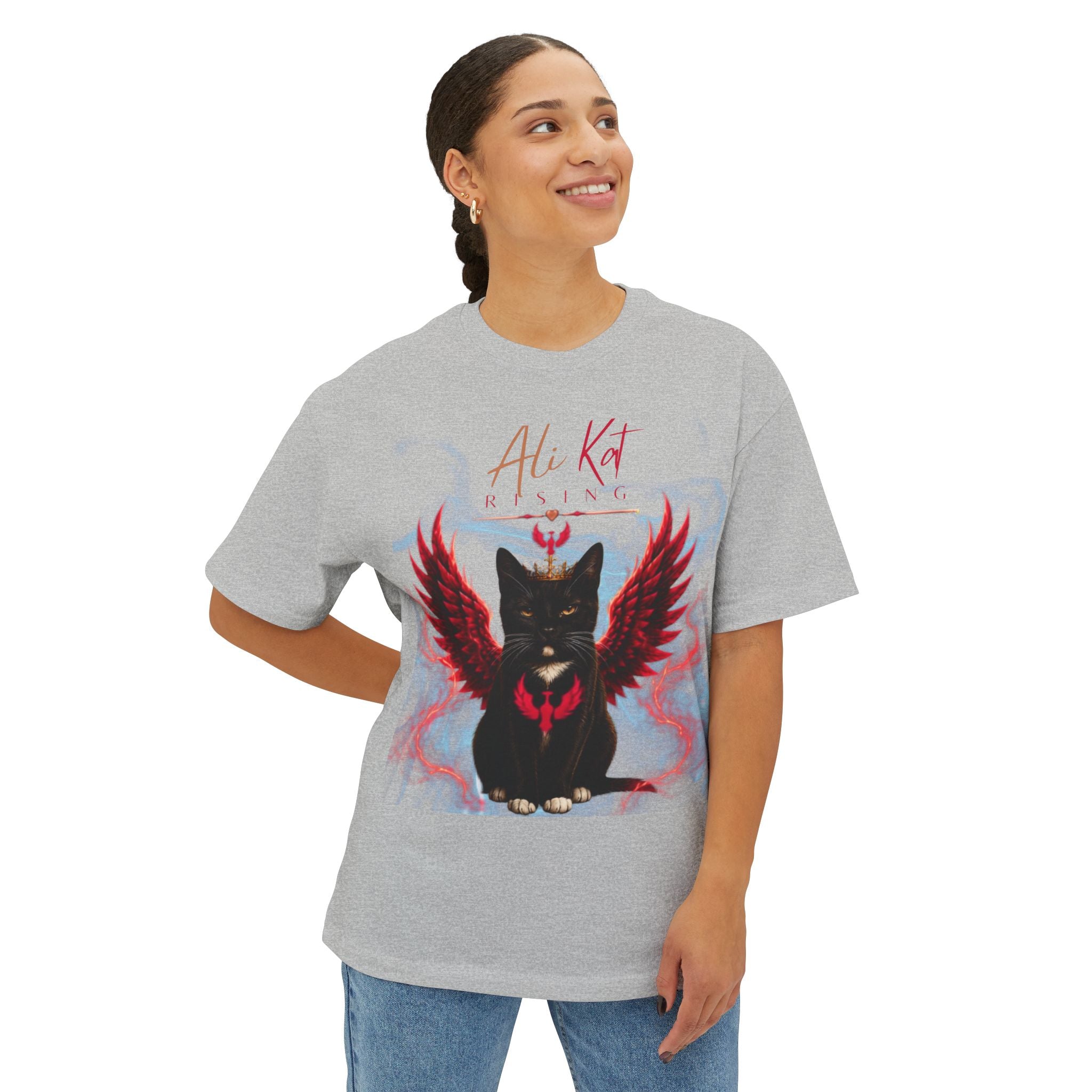 A smiling person stands against a white background, wearing the AliKat Rising Phoenix Season: Vol. 1 Signature Oversized Tee—an oversized unisex t-shirt in gray premium heavyweight cotton featuring a black cat graphic with red wings and the text “Ali Kat Rising.”.