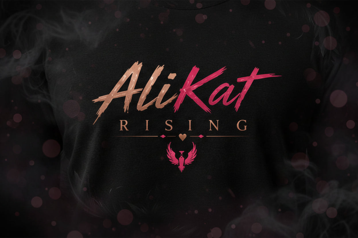 A black shirt with the text AliKat RISING in bold brush font, with a pink phoenix logo, heart, and dots below the text. Smoky, pinkish effects surround the design.