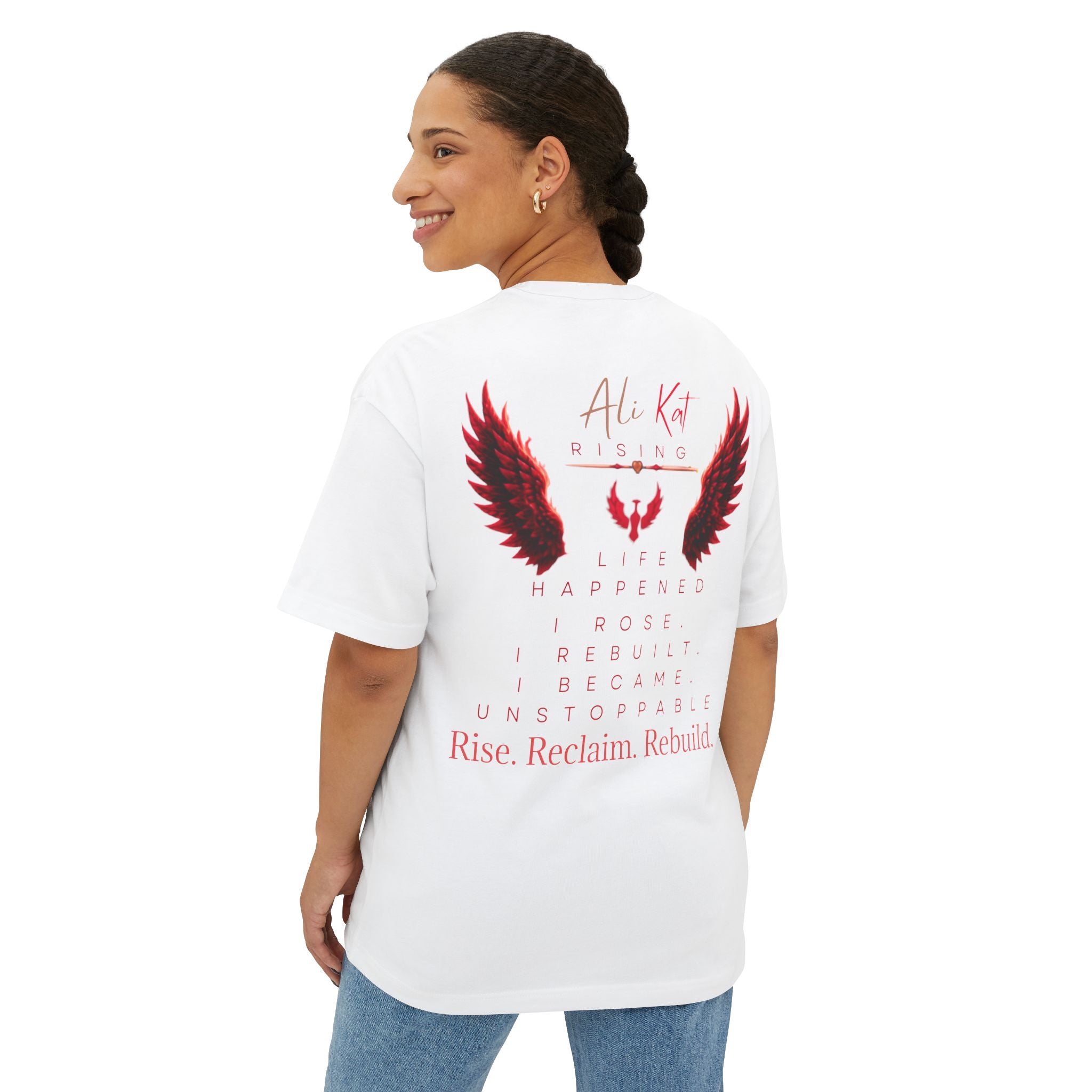 A woman with braided hair wears the AliKat Rising Phoenix Season: Vol. 1 Signature Oversized Tee—a unisex T-shirt crafted from premium heavyweight cotton, featuring bold red and black wing graphics and motivational text on the back. She stands sideways, smiling as she looks over her shoulder.