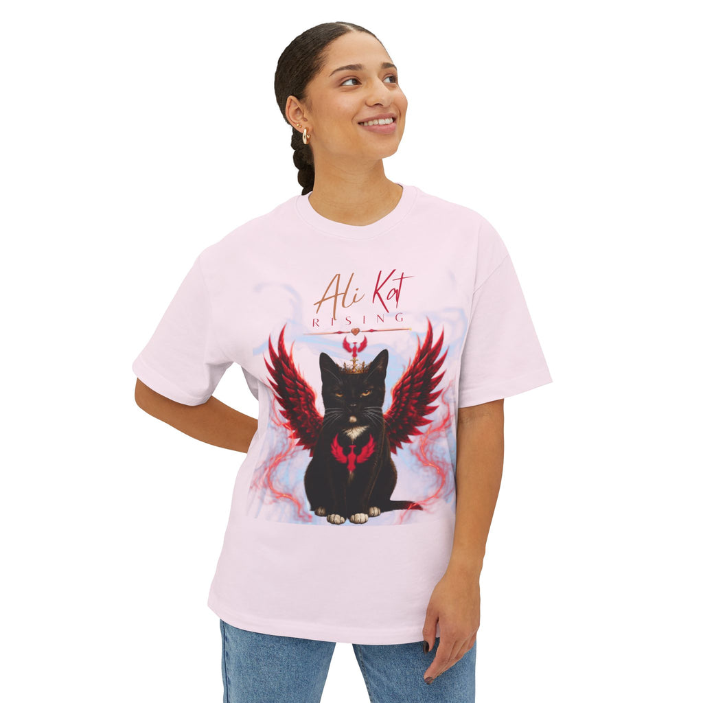 A smiling person wears the AliKat Rising Phoenix Season: Vol. 1 Signature Oversized Tee—a premium, heavyweight cotton, oversized unisex t-shirt in light pink, featuring a graphic of a black cat with red wings and a crown, along with the text "Ali Kat Rising" above the design.