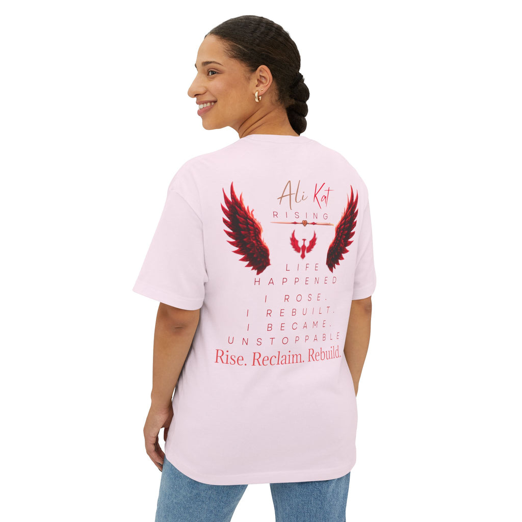 A woman is smiling and looking over her shoulder while wearing the AliKat Rising Phoenix Season: Vol. 1 Signature Oversized Tee in light pink. Crafted from premium heavyweight cotton, this unisex t-shirt features a striking phoenix graphic and inspirational text—including “Rise. Reclaim. Rebuild.”—on the back.