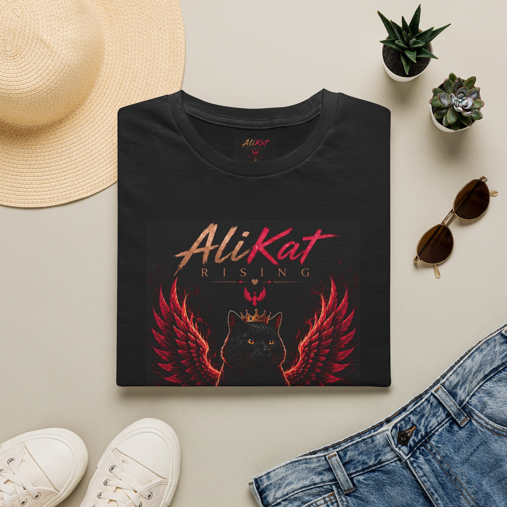 A folded Phoenix Vol. 1 Graphic Tee by AliKat Rising is artfully arranged on a beige surface, surrounded by a hat, plants, sunglasses, and denim jeans. Available in sizes S through 2XL, this black T-shirt features a striking design of a black cat with red wings and the “AliKat Rising” text.