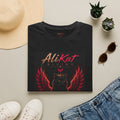 A folded Phoenix Vol. 1 Graphic Tee by AliKat Rising is artfully arranged on a beige surface, surrounded by a hat, plants, sunglasses, and denim jeans. Available in sizes S through 2XL, this black T-shirt features a striking design of a black cat with red wings and the “AliKat Rising” text.