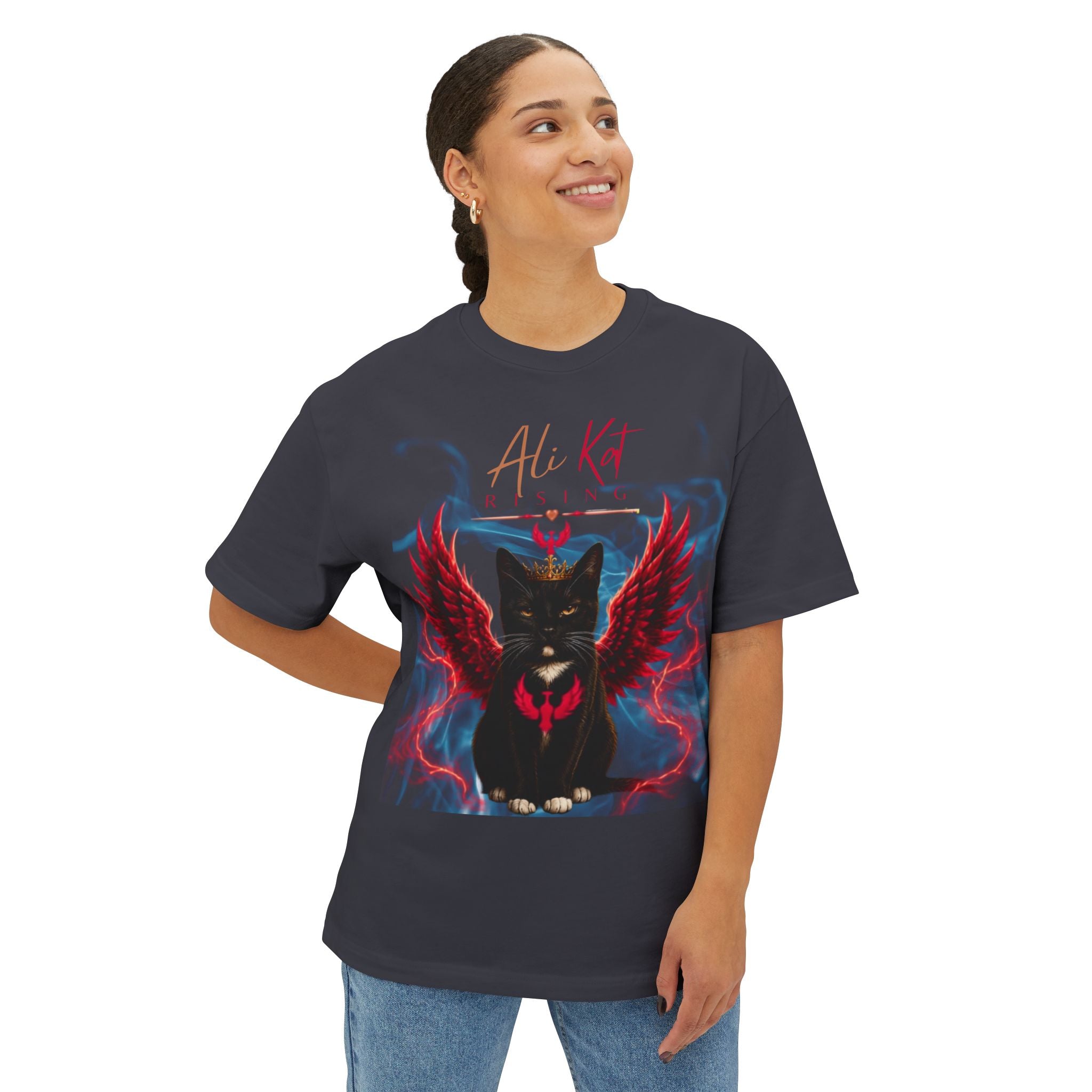 Wearing the AliKat Rising Phoenix Season: Vol. 1 Signature Oversized Tee—a dark, premium heavyweight cotton unisex shirt adorned with a bold graphic of a black cat with red wings and blue lightning, plus the text "Ali Kat RISING"—the person is smiling and looking slightly to the side.