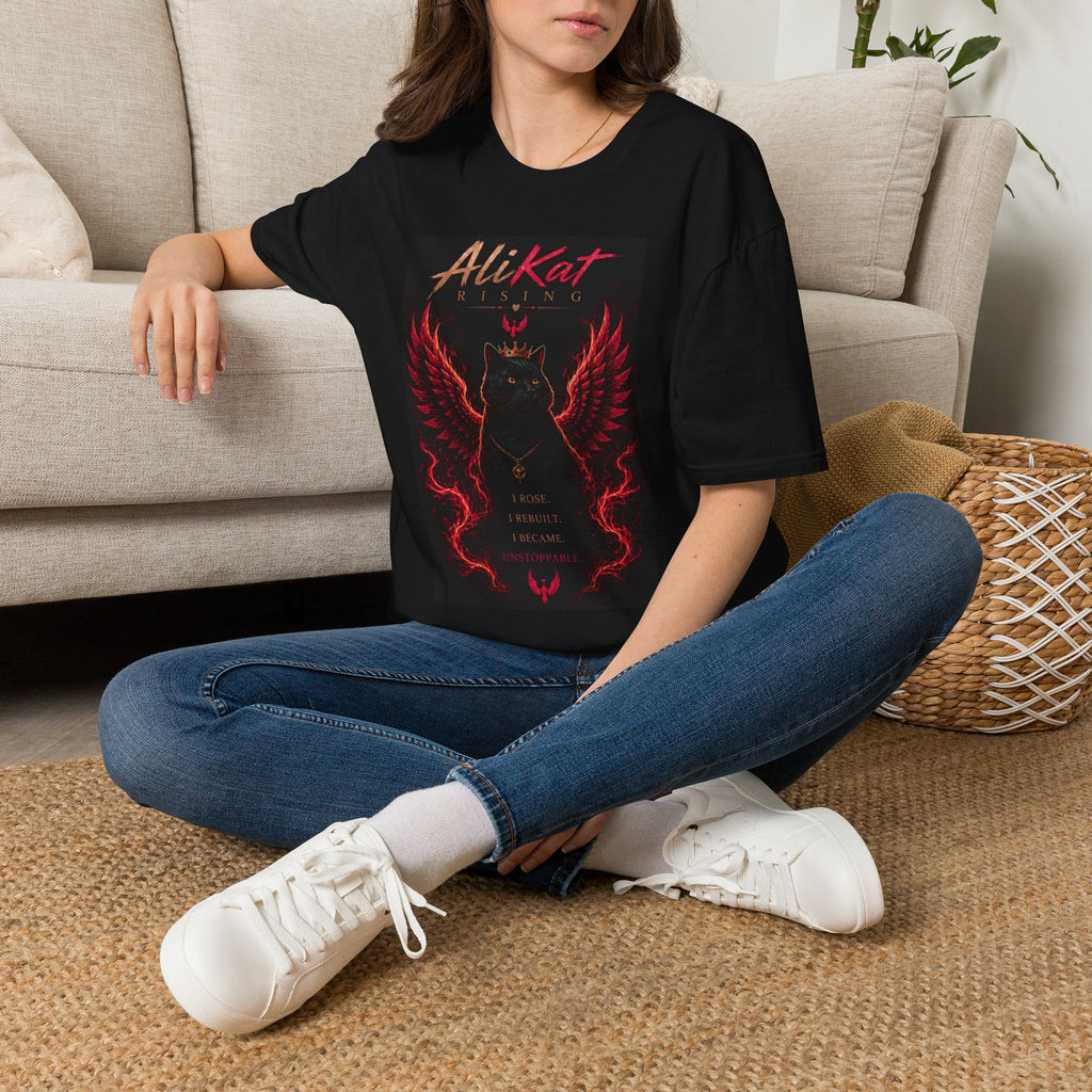 A woman sits cross-legged on the floor, wearing the Phoenix Vol. 1 Graphic Tee by AliKat Rising—a black t-shirt featuring a striking red cat with wings and the empowering text: "I roar. I resist. I become. Unstoppable." Available in sizes S, M, L, XL, and 2XL, this bold AliKat Rising design is perfect for anyone ready to make a statement.